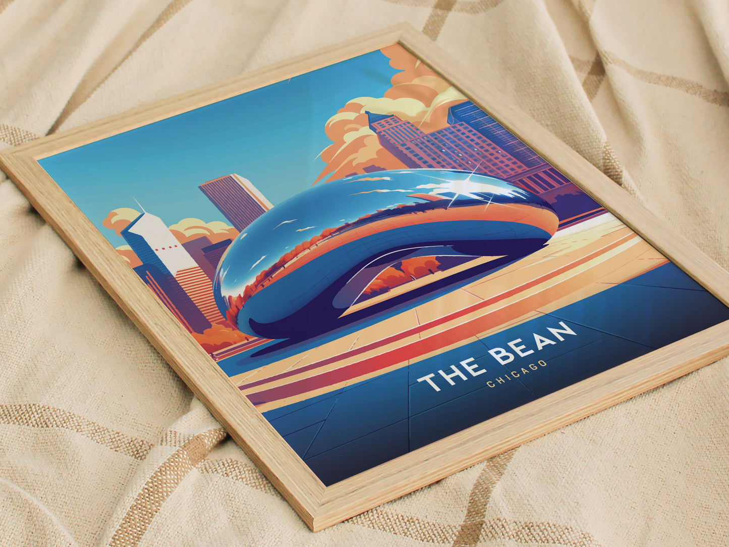 A framed poster of The Bean in Chicago, featuring Anish Kapoors Cloud Gate with a vibrant city skyline and bright blue sky, rests on a textured fabric.