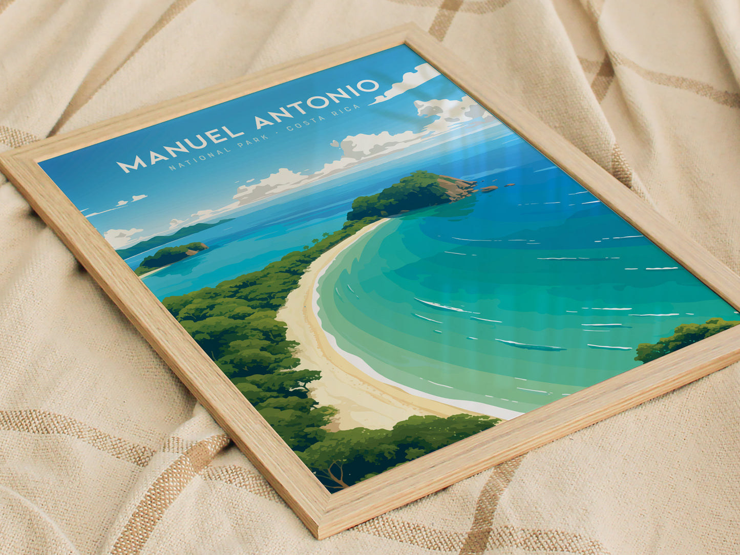 A framed poster of Manuel Antonio National Park rests on a fabric surface, depicting a vibrant coastal scene with turquoise waters, white sandy beaches, and lush green foliage.
