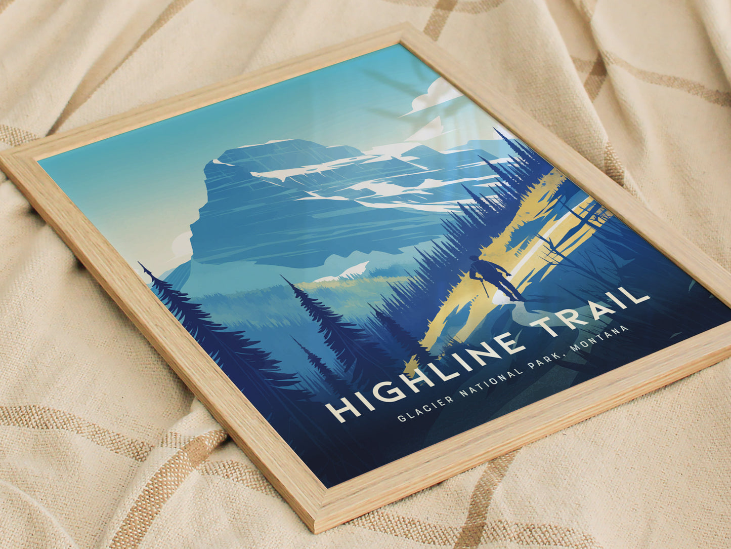 A framed poster of the Highline Trail in Glacier National Park, featuring a hiker silhouette against a backdrop of mountains and trees, rests on a textured beige fabric.