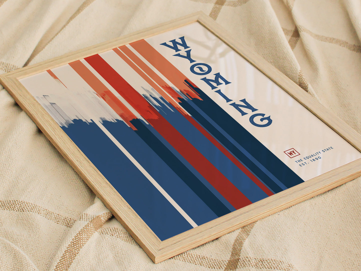 Framed Wyoming poster featuring bold red, white, and blue vertical stripes with stylized text on a beige background, resting on a textured fabric surface.