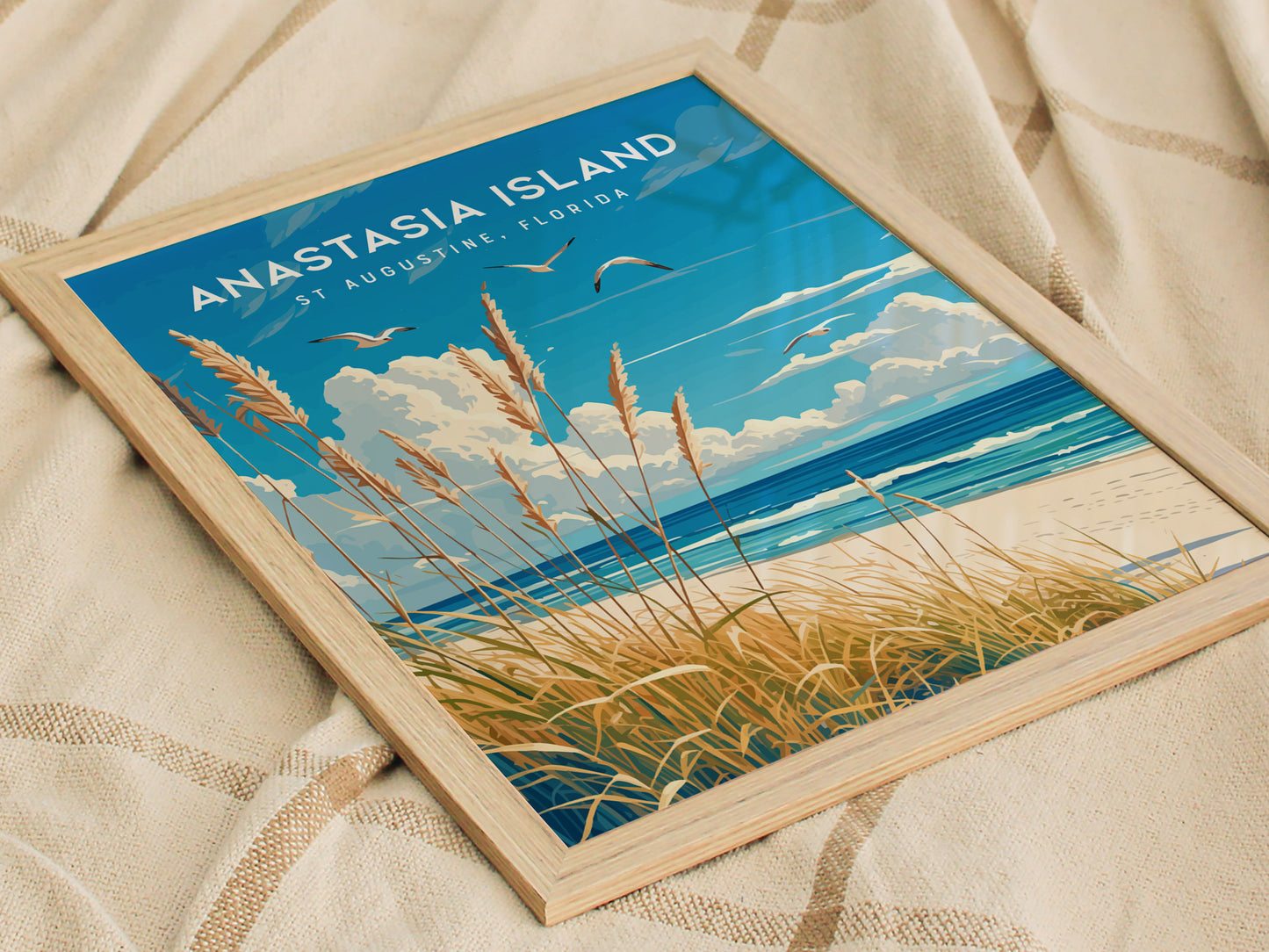 Framed artwork showcasing Anastasia Island, featuring sea oats, golden dunes, and seabirds over a tranquil ocean. The poster is placed on a textured beige fabric background.