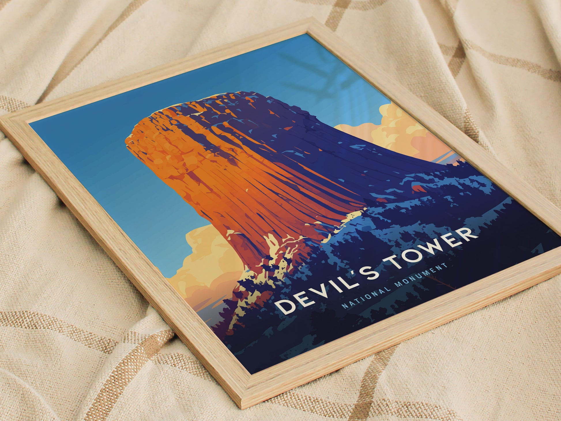 A framed poster of Devils Tower with vibrant blue and orange hues rests on a soft, textured beige fabric. The text reads Devils Tower National Monument.