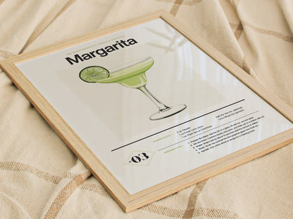 Framed margarita cocktail poster with a lime illustration, resting on a light beige fabric. The artwork features a recipe and elegant typography.