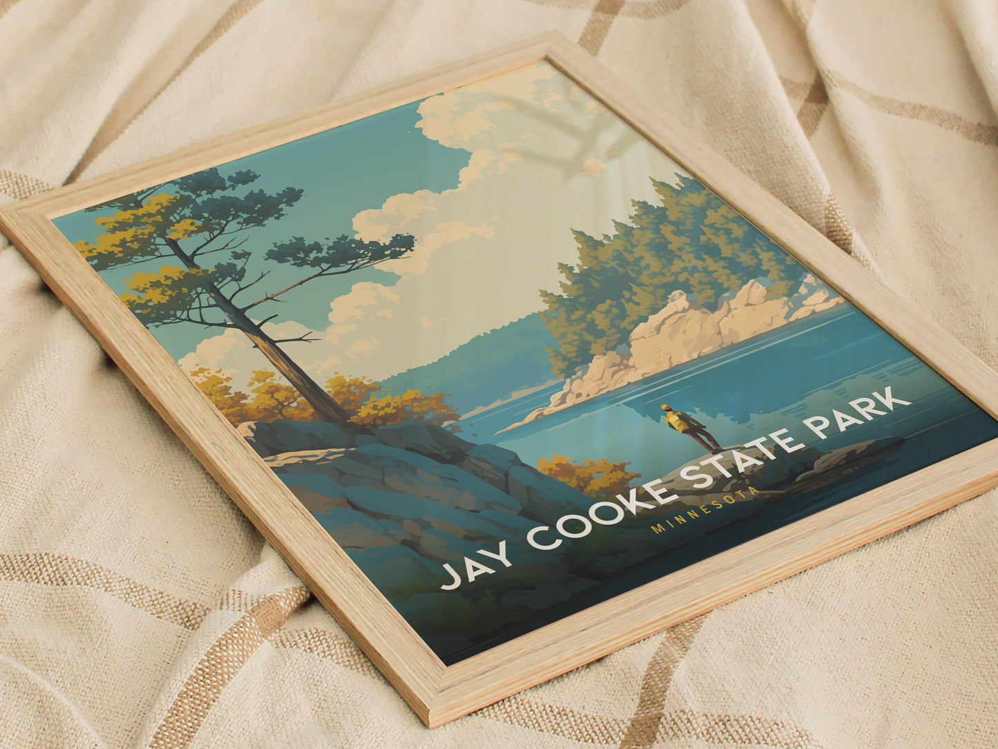 Framed Jay Cooke State Park poster resting on a patterned fabric, featuring a scenic landscape with trees, rocks, and a river under a blue sky.