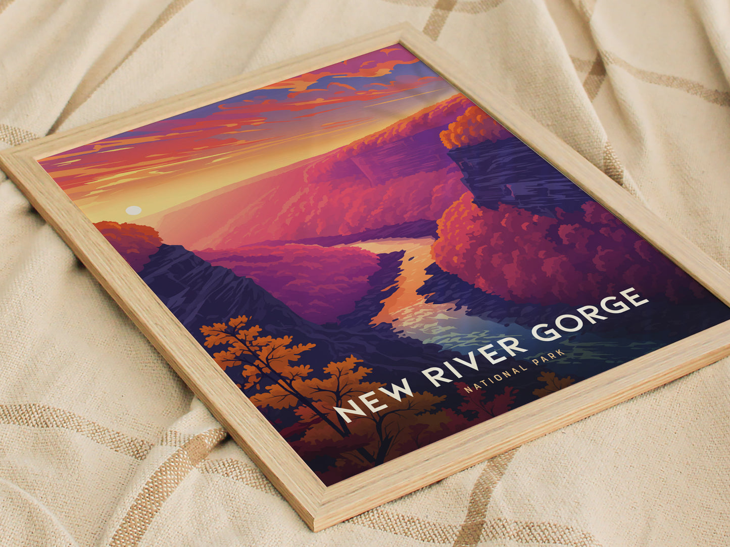 A framed poster of New River Gorge National Park features a vibrant sunset over a winding river and colorful, forested canyon. The artwork rests on a textured fabric surface.