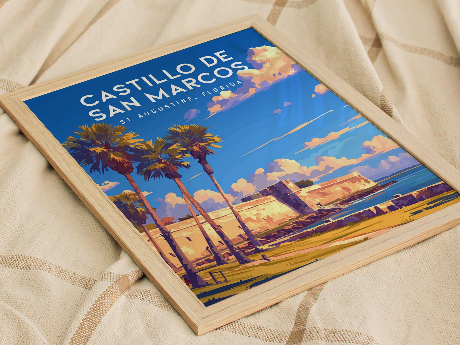 Framed poster depicting Castillo de San Marcos with palm trees under a vibrant blue sky and scattered clouds, resting on a textured fabric surface.