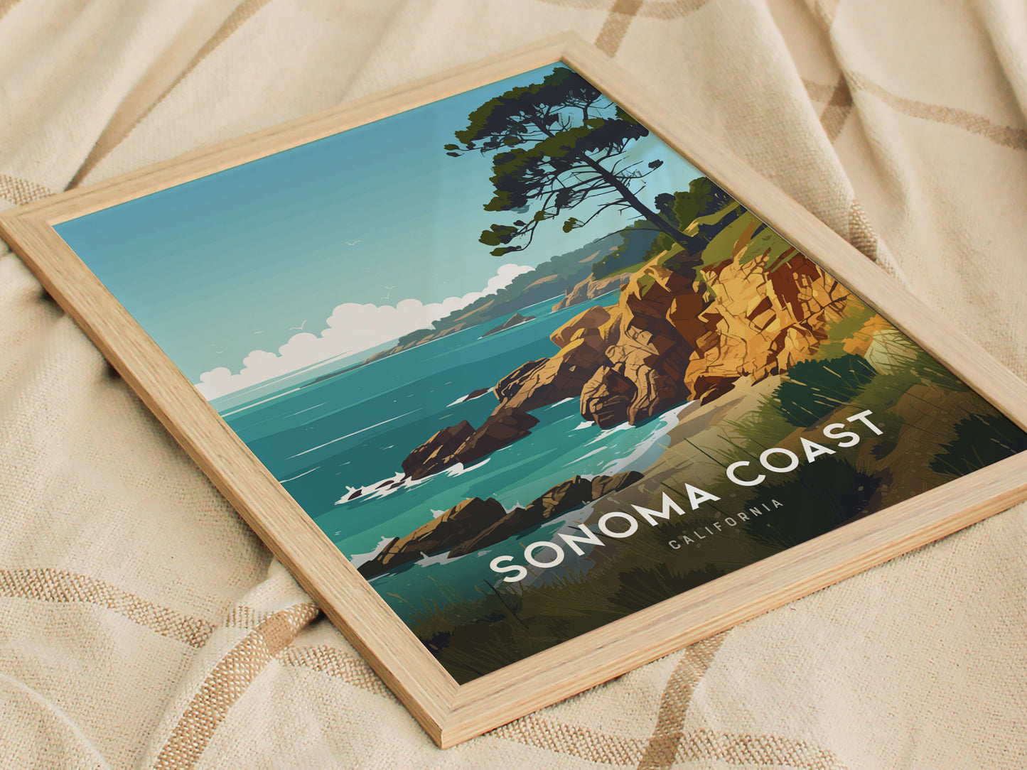 Framed poster of Sonoma Coast featuring rocky cliffs, a tree, and the ocean under a blue sky, resting on a textured beige fabric.