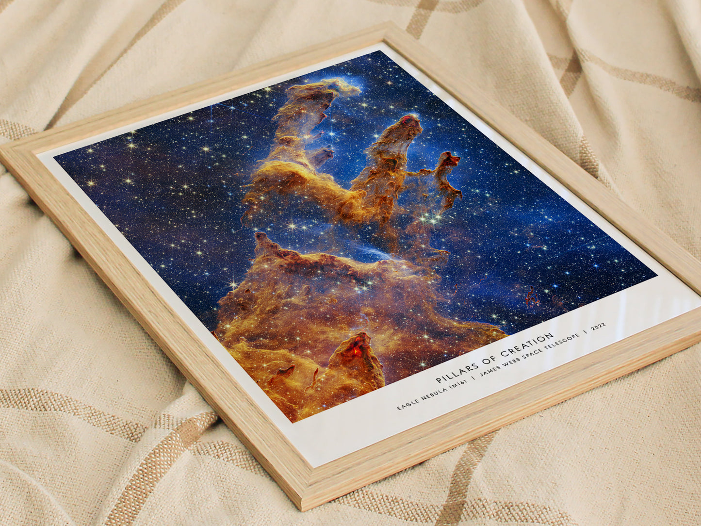 Framed Pillars of Creation poster placed on a cozy fabric surface, highlighting the blue starfield and orange nebula formations.