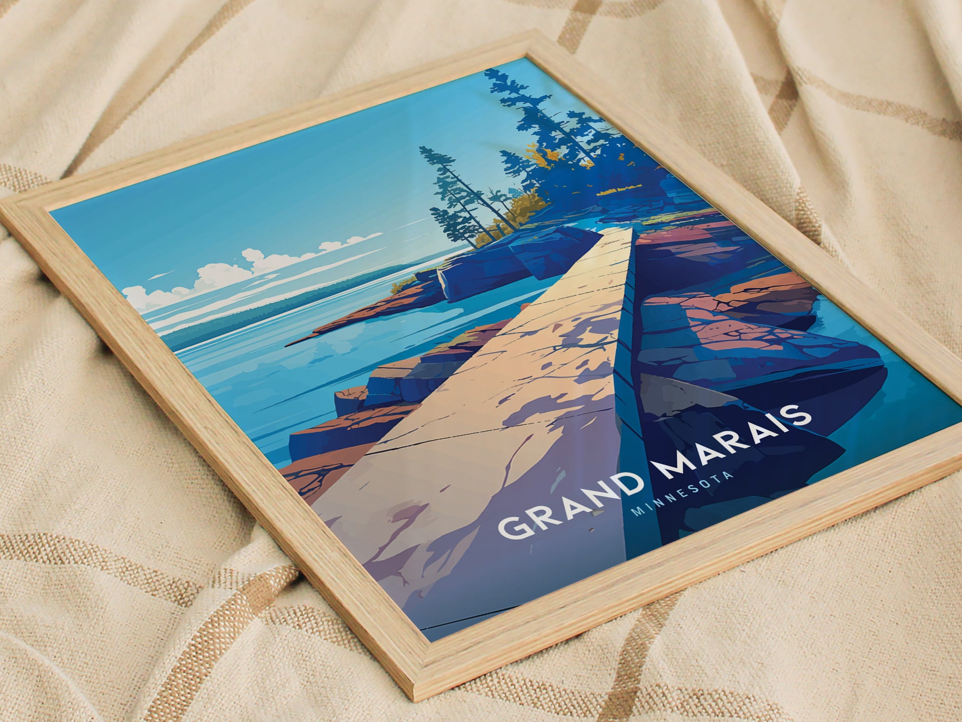 Framed poster featuring a colorful illustration of Artist’s Point in Grand Marais, Minnesota, showcasing rocky shores and a vibrant blue sky. The poster rests on a beige fabric background.