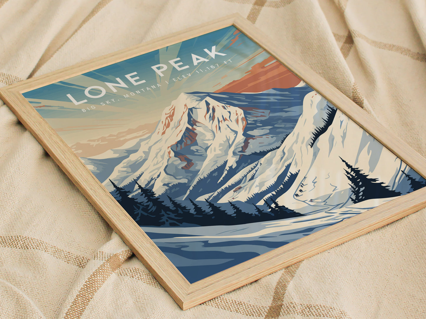 Framed poster featuring Lone Peak in Big Sky, Montana, with a stylized depiction of the snowy mountain and pine trees, resting on a textured beige fabric.