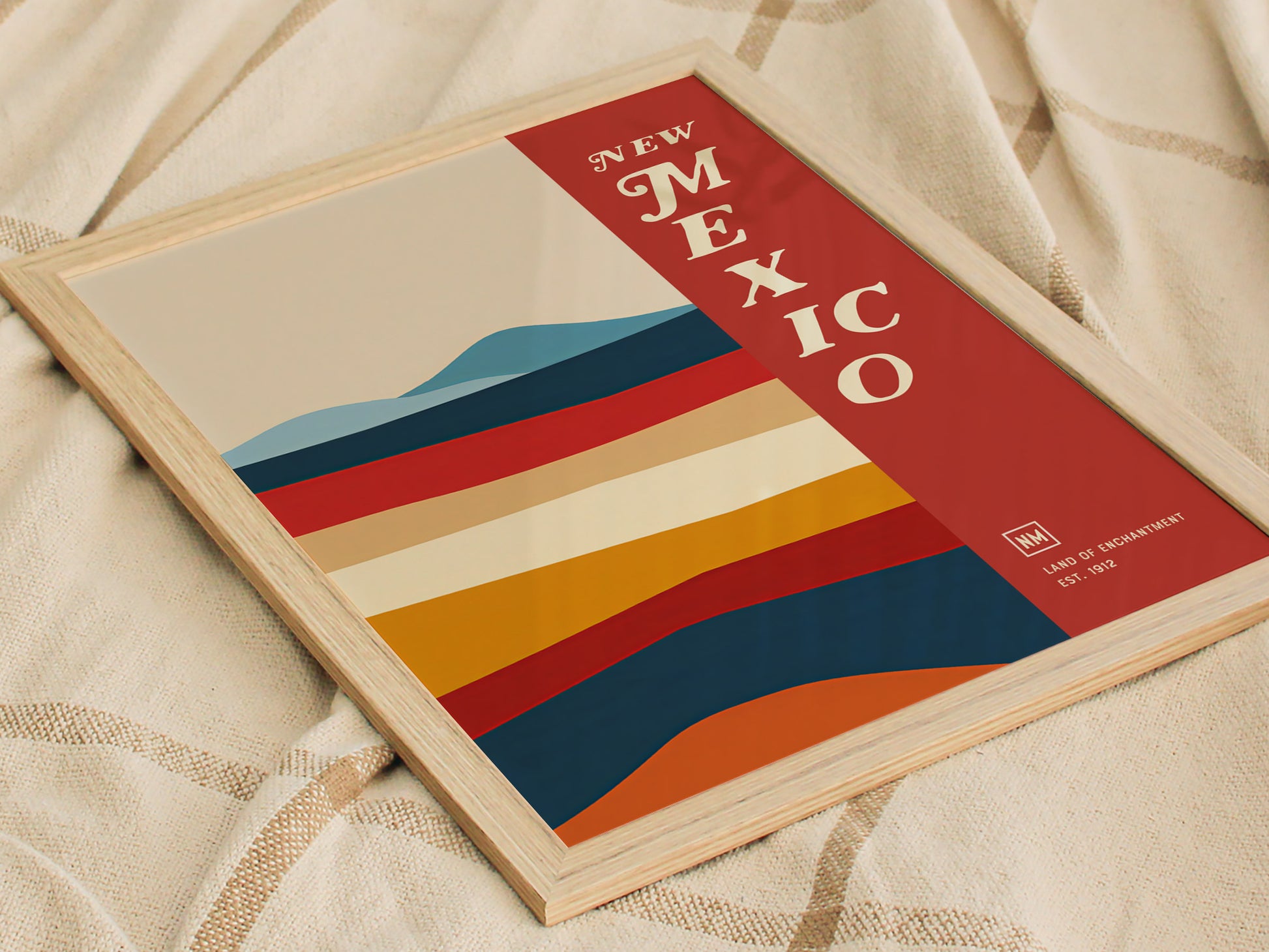 A framed poster titled New Mexico features colorful abstract desert landscapes with layered horizontal stripes in warm tones, set against a beige background. It rests on a textured fabric.