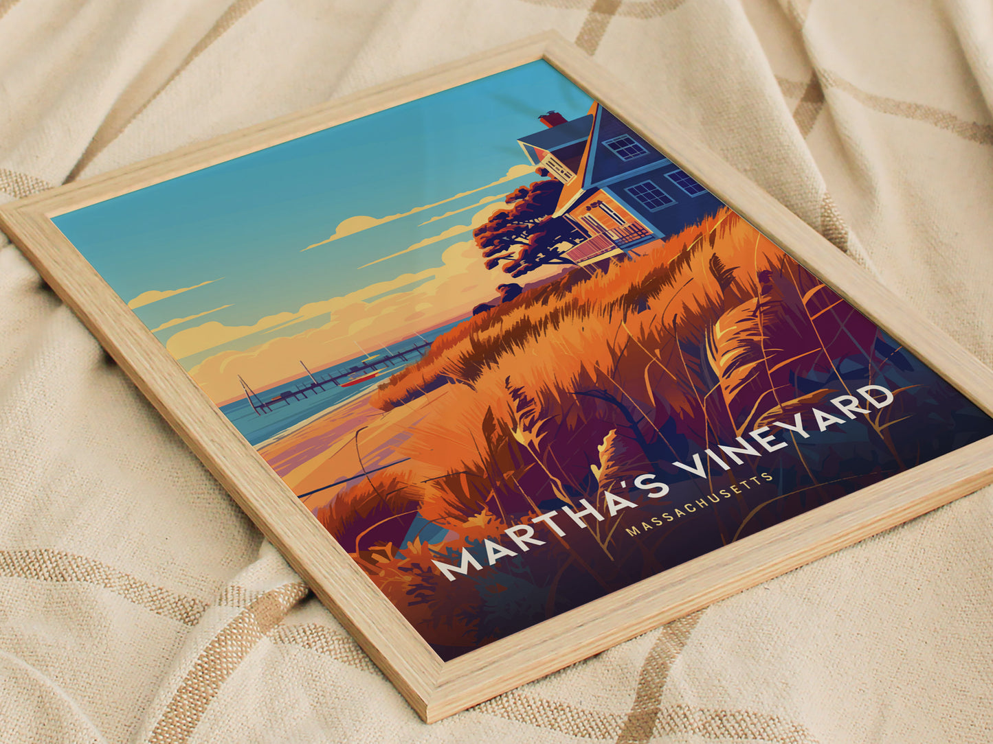 A framed poster of Marthas Vineyard, Massachusetts, features a coastal scene with a house, orange grasses, and a blue sky. The artwork rests on a textured beige fabric.