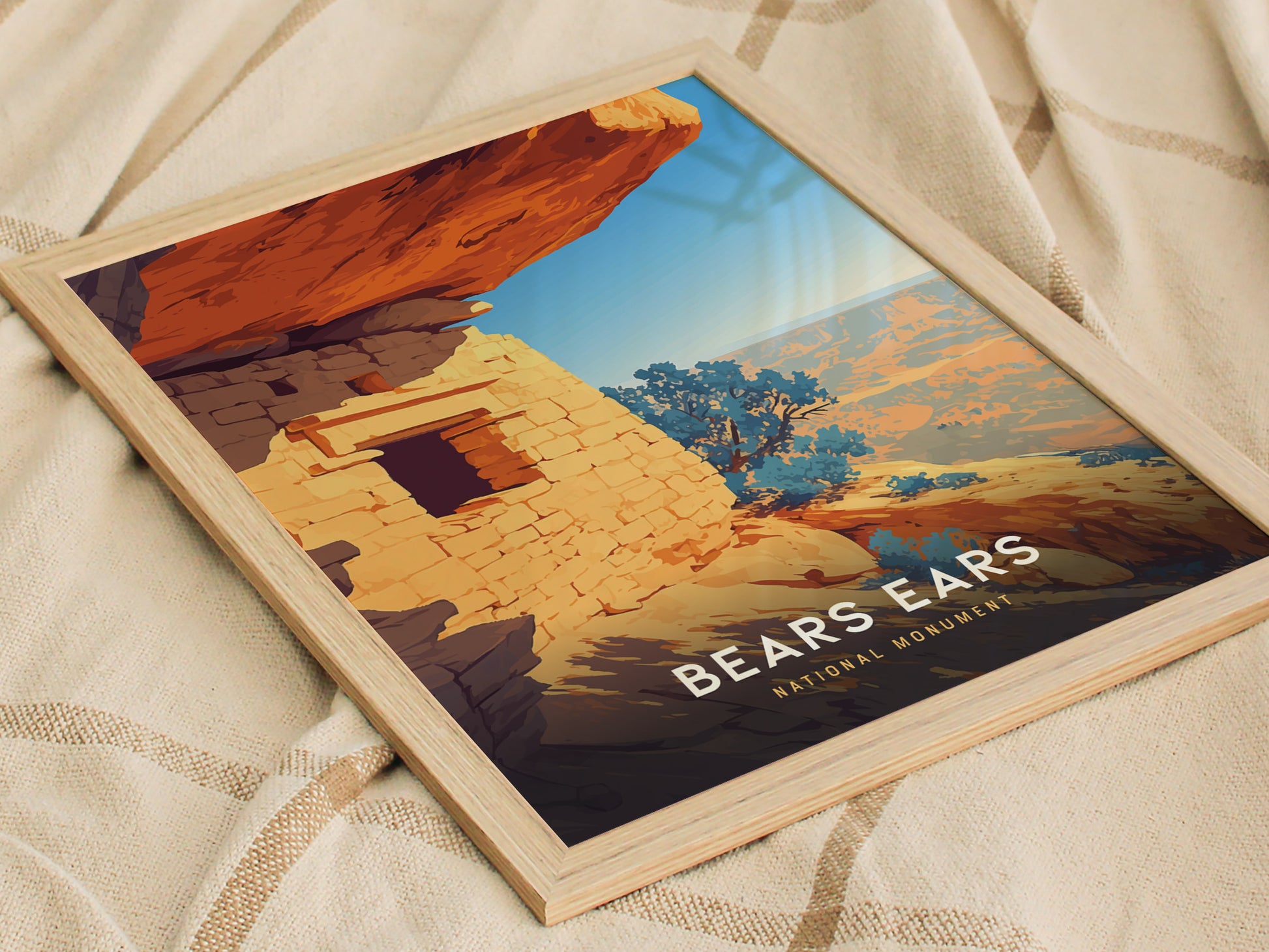 A framed poster of Bears Ears National Monument displays a vibrant illustration of sandstone cliffs and ruins under a blue sky, resting on a beige fabric surface.