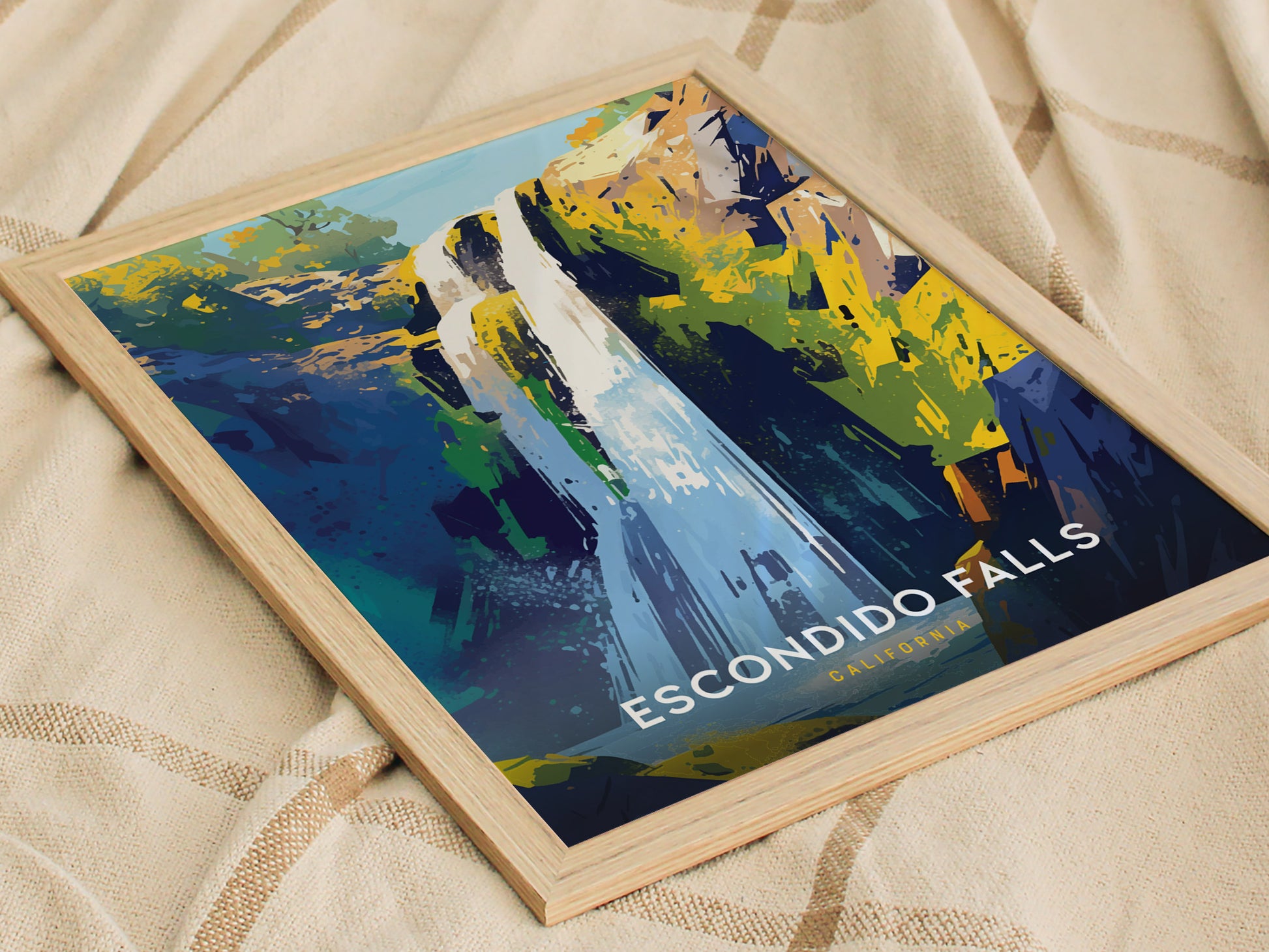 A framed art print of Escondido Falls features a stylized depiction of a waterfall with vibrant blues, greens, and yellows. Its placed on a textured beige fabric.