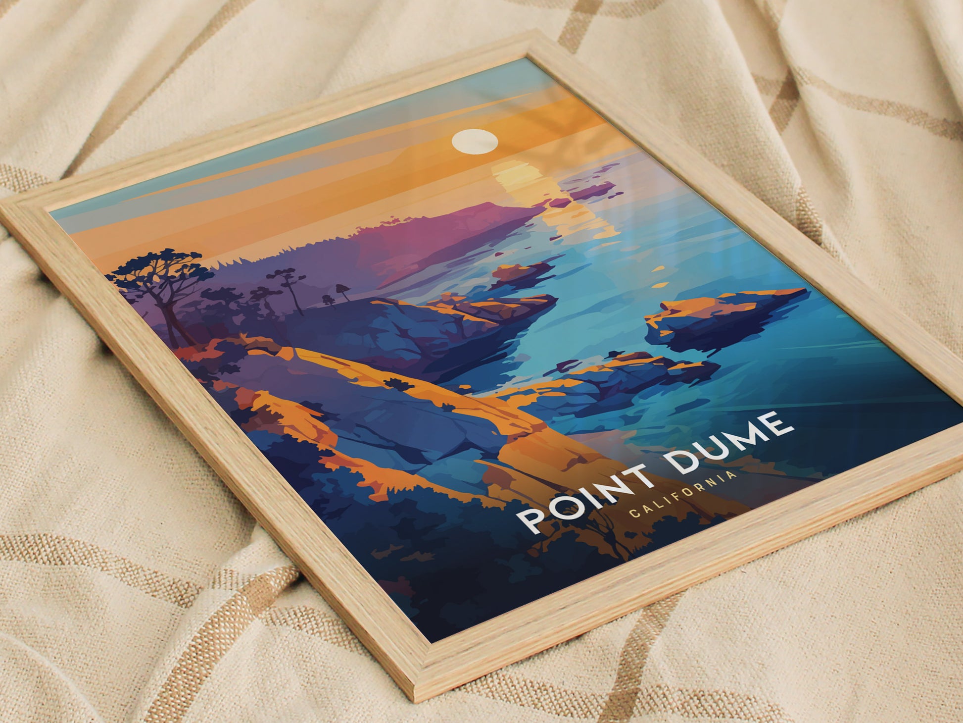 Framed poster of Point Dume, California, showcasing vibrant cliffs and the Pacific Ocean under a setting sun, resting on a beige fabric background.