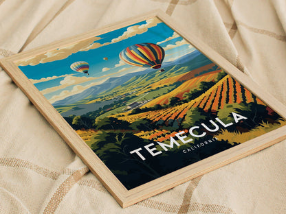 A vibrant poster of Temecula, California, features colorful hot air balloons floating over scenic vineyards and rolling hills, framed in light wood, resting on a textured fabric.