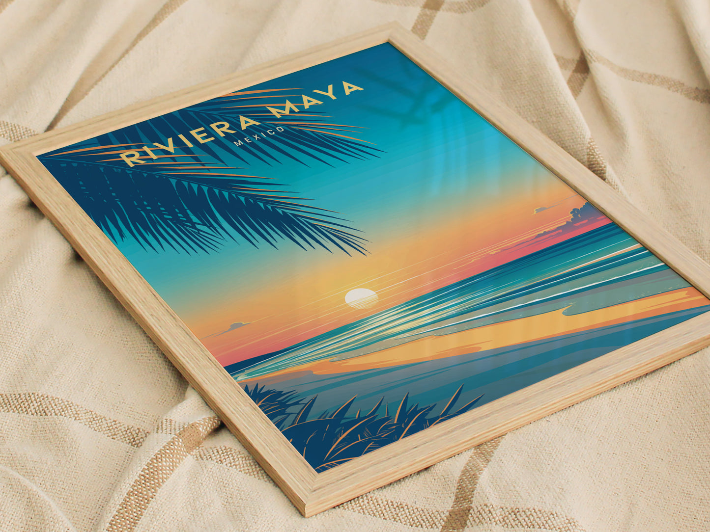 Framed travel poster of Riviera Maya, Mexico, featuring a vibrant sunset over the ocean with palm leaves in the foreground, placed on a textured beige fabric.