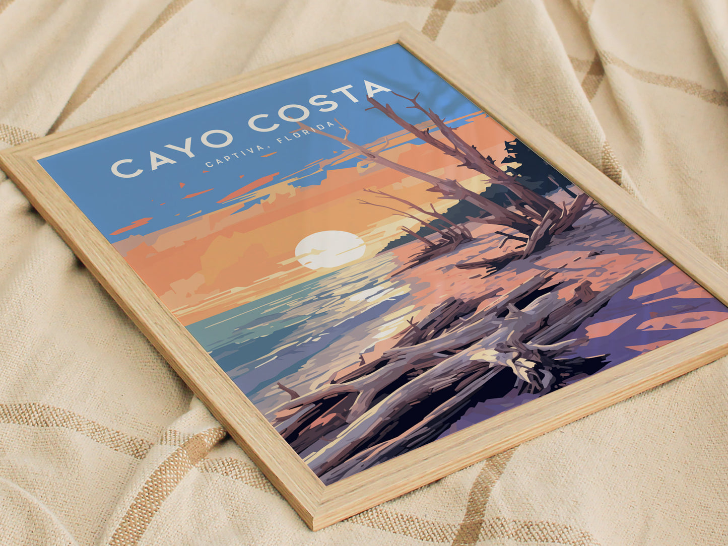 A poster depicting a sunset over a beach with driftwood in the foreground, labeled Cayo Costa, Captiva Florida. The artwork is framed and placed on a textured fabric surface.