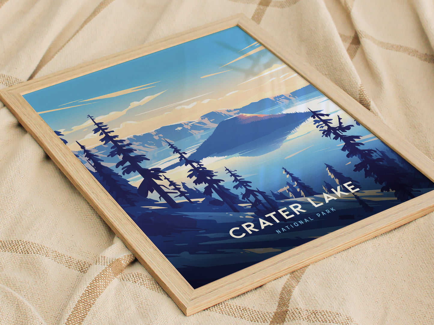 Framed poster of Crater Lake National Park features a stylized landscape with deep blue water, surrounding mountains, and silhouetted trees, resting on a textured beige fabric.
