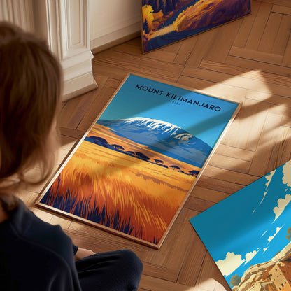 A person looks at a colorful Mount Kilimanjaro poster on a wooden floor, featuring a snow-capped peak and savanna landscape. Additional artwork is visible nearby.