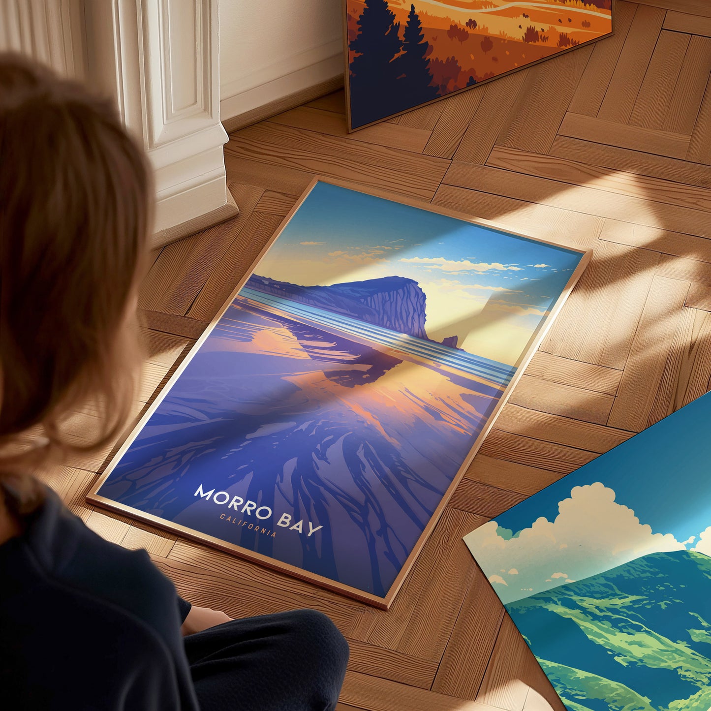 A person sits on a wooden floor, looking at a colorful poster of Morro Bay featuring Morro Rock and a vivid sunset. Nearby, other landscape posters are partially visible.