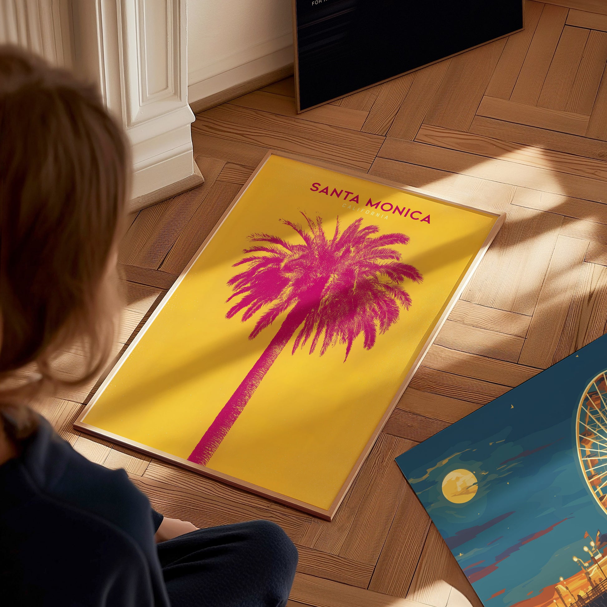 A person looks at a vibrant Santa Monica poster featuring a pink palm tree on a yellow background. Another colorful print, partially visible, lies nearby on a wooden floor.