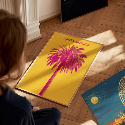 A person looks at a vibrant Santa Monica poster featuring a pink palm tree on a yellow background. Another colorful print, partially visible, lies nearby on a wooden floor.