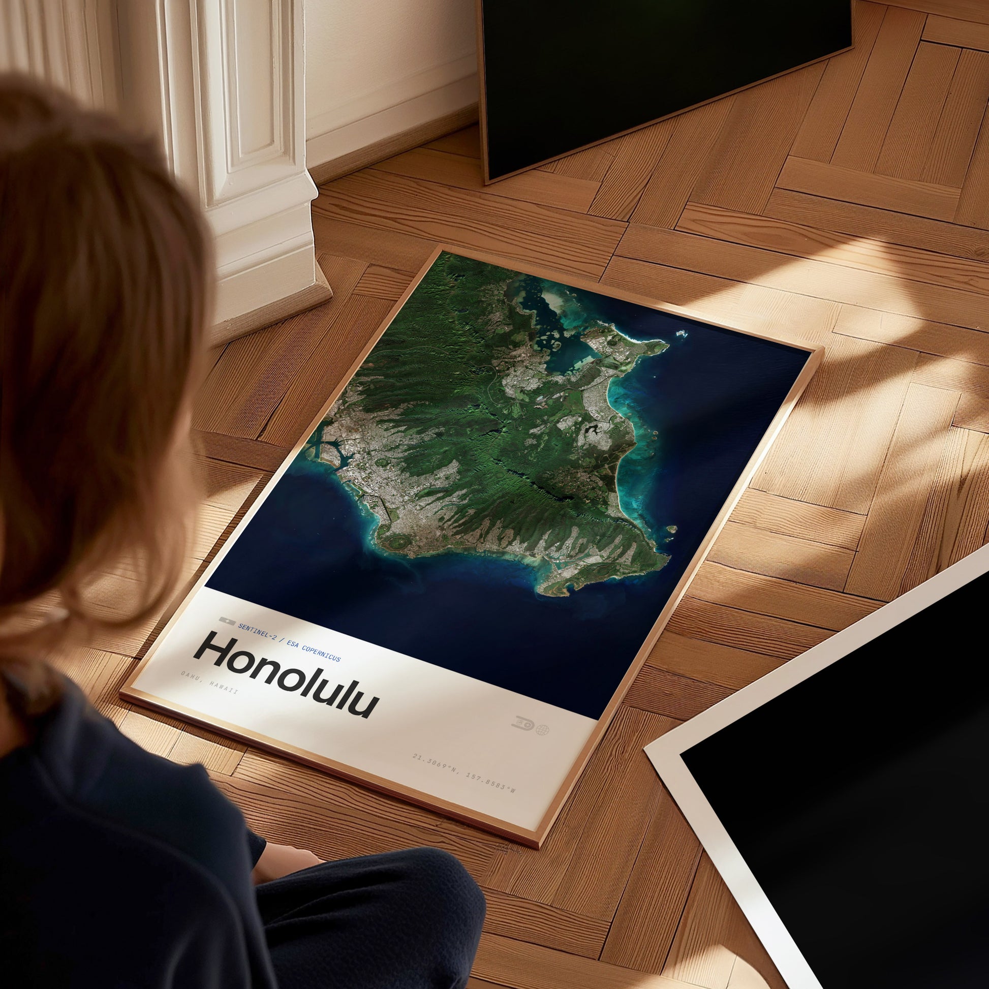 A person sits on a wooden floor, gazing at a framed travel print of Honolulu. The artwork features a detailed aerial view of the island, showcasing its lush landscapes, coastlines, and urban areas beneath a bright blue ocean.
