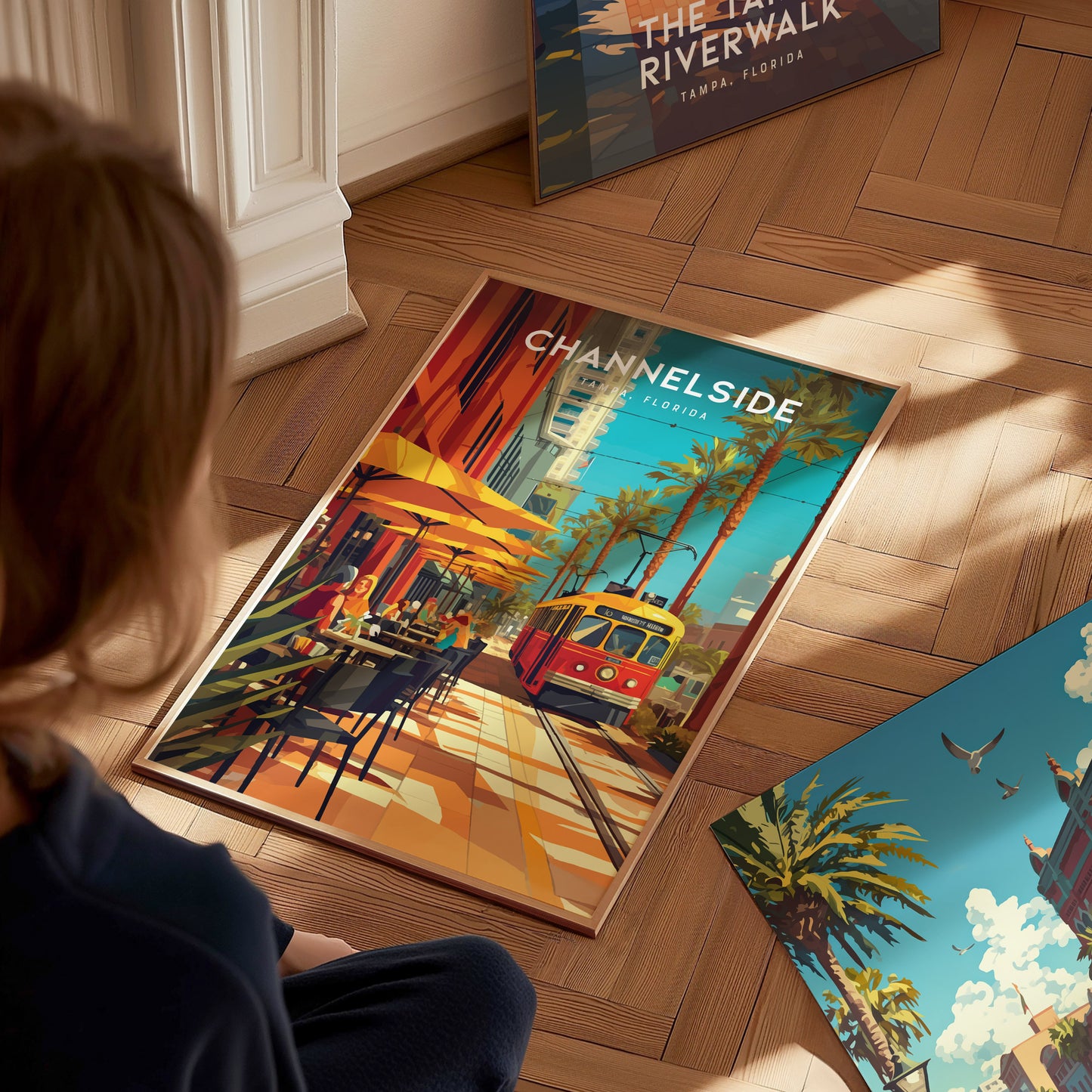 A person looks at a colorful framed poster of Channelside, Tampa, featuring a vibrant street scene with a yellow and red trolley, palm trees, and outdoor dining.
