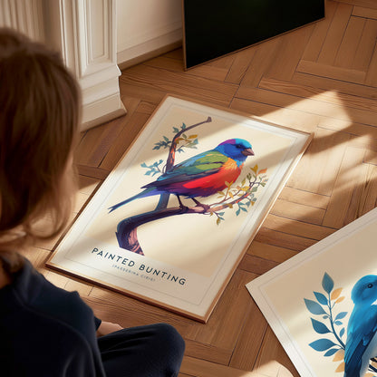 A child sits on a wooden floor, looking at a colorful art print of a Painted Bunting perched on a branch. The vibrant illustration features shades of blue, yellow, and red, surrounded by delicate leaves, while another print lies nearby.