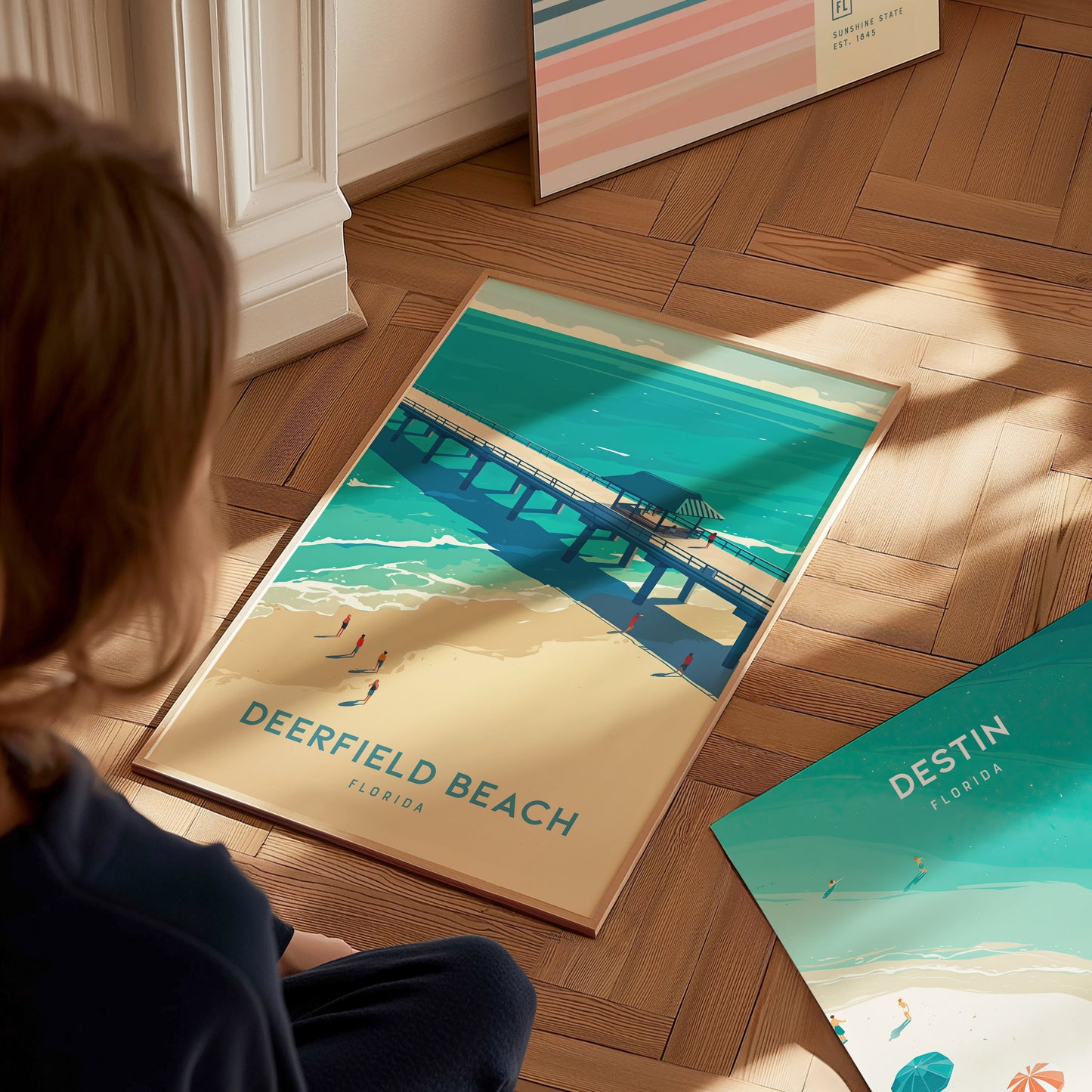 A person looks at a Deerfield Beach poster on a wooden floor, depicting a pier over turquoise waters and golden sands under a sunny sky.