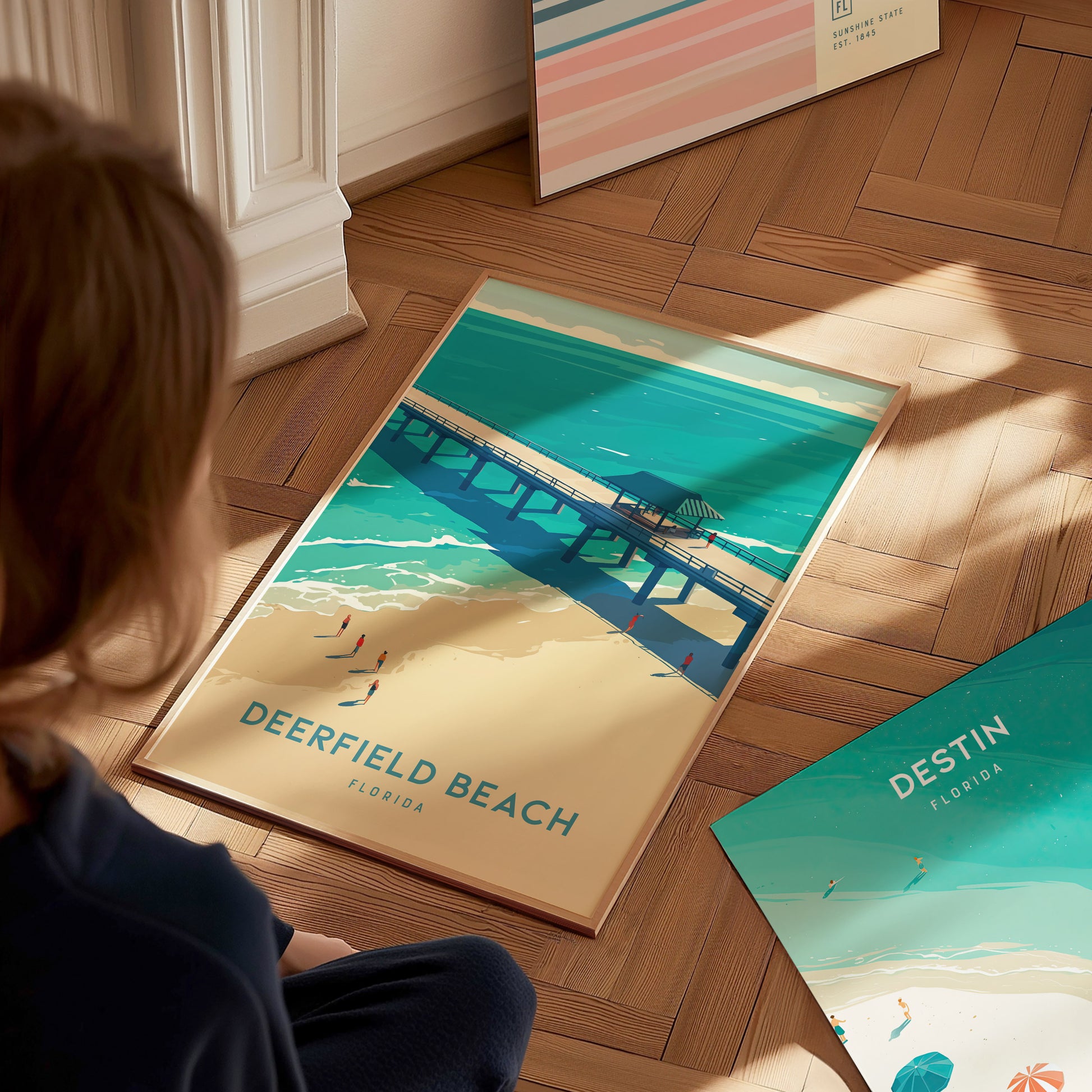 A person looks at a Deerfield Beach poster on a wooden floor, depicting a pier over turquoise waters and golden sands under a sunny sky.
