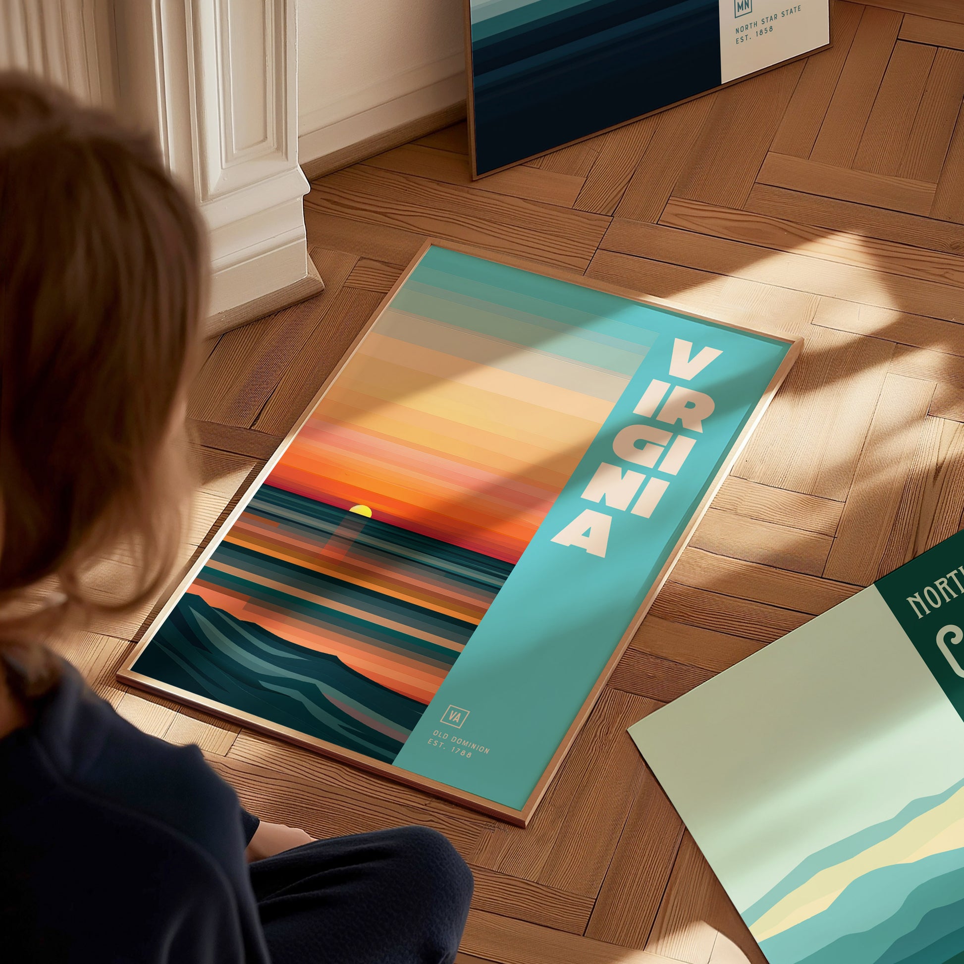 A person sits on a wooden floor, looking at a colorful poster titled Virginia with abstract horizontal stripes and a sunset design. Other posters are partially visible nearby.