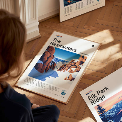 A person sits on a wooden floor, viewing a framed poster titled The Headwaters featuring a snowy mountain scene with rocky ridgelines. Additional posters are partially visible nearby.