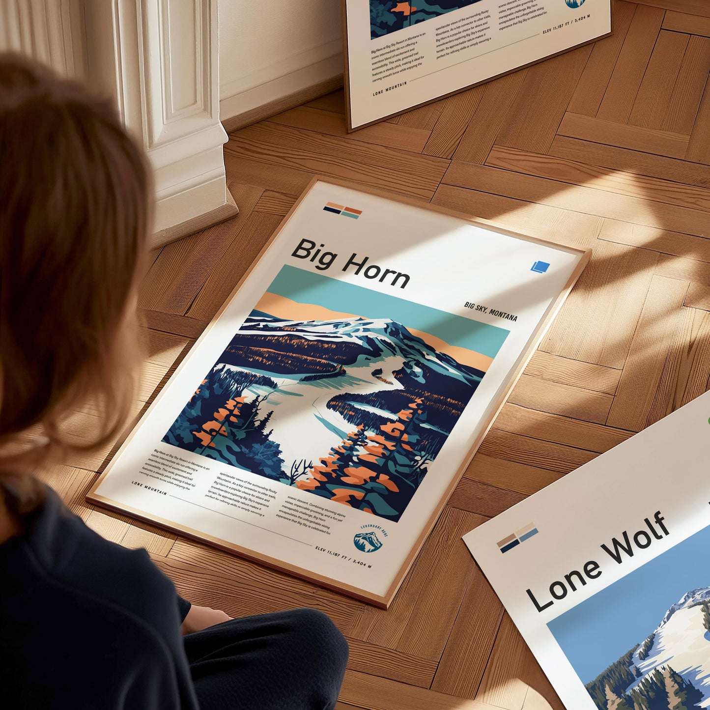 A person sits on the floor looking at a framed poster of Big Horn ski run, featuring a stylized mountain landscape. Other posters are partially visible on the wooden floor.