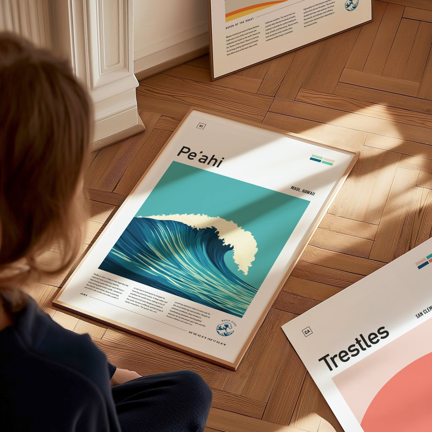 A person sits on a wooden floor viewing a framed Peahi surf poster depicting a large wave. Sunlight casts shadows on nearby surfaces, creating a warm ambiance.
