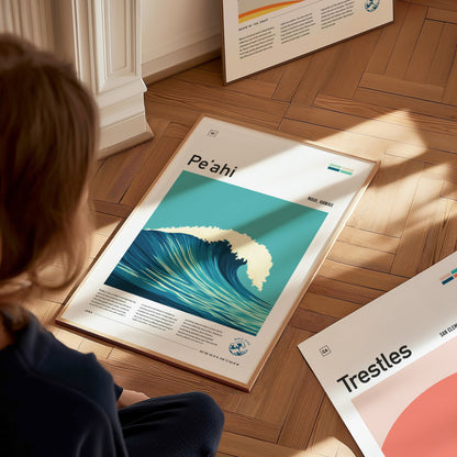 A person sits on a wooden floor viewing a framed Peahi surf poster depicting a large wave. Sunlight casts shadows on nearby surfaces, creating a warm ambiance.