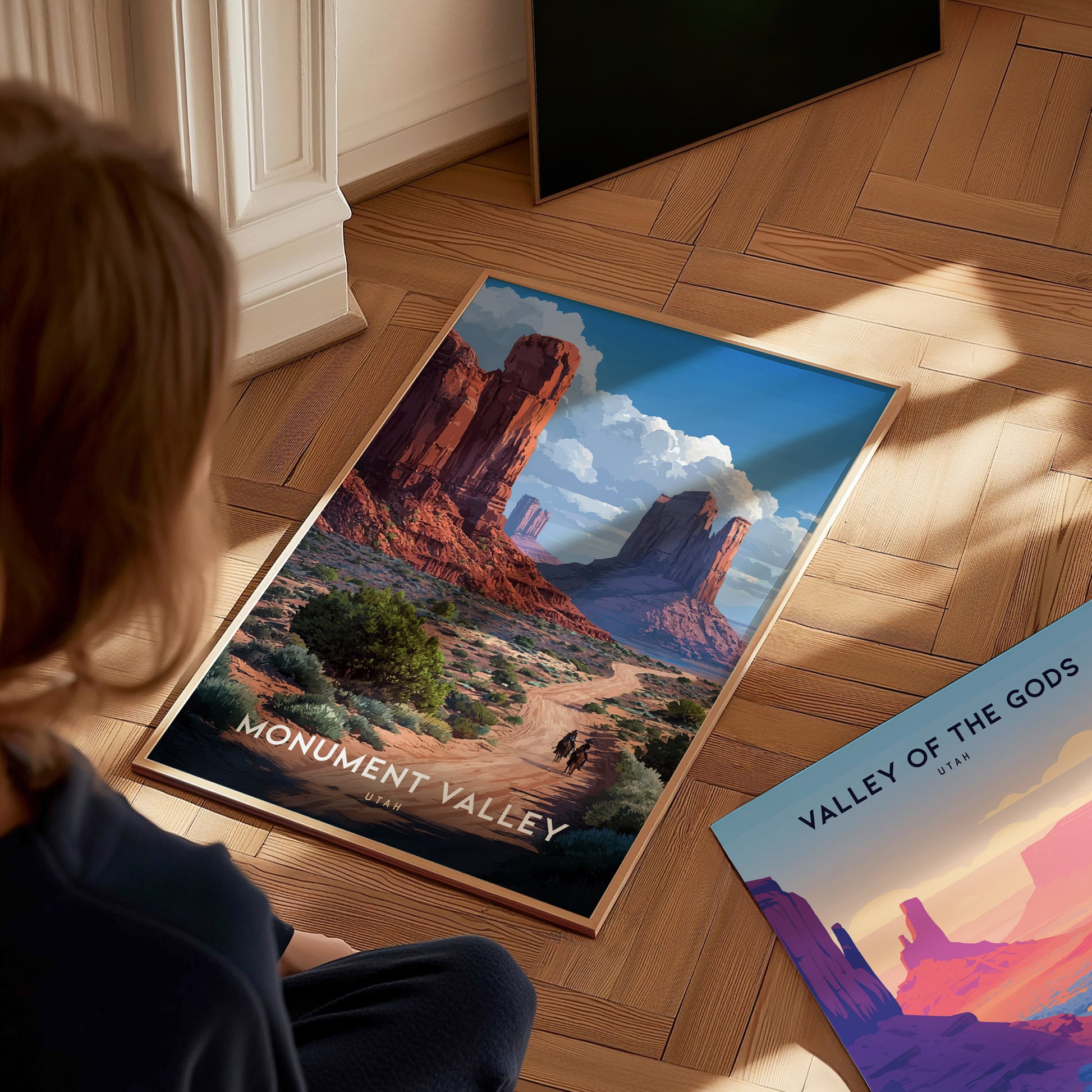 A person views a Monument Valley poster on a wooden floor, depicting red sandstone mesas and a desert landscape under a blue sky.