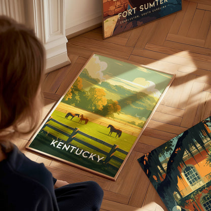 A person views a Kentucky poster featuring horses, green pastures, and a black fence, lying on a wooden floor. Nearby, other art prints are visible.