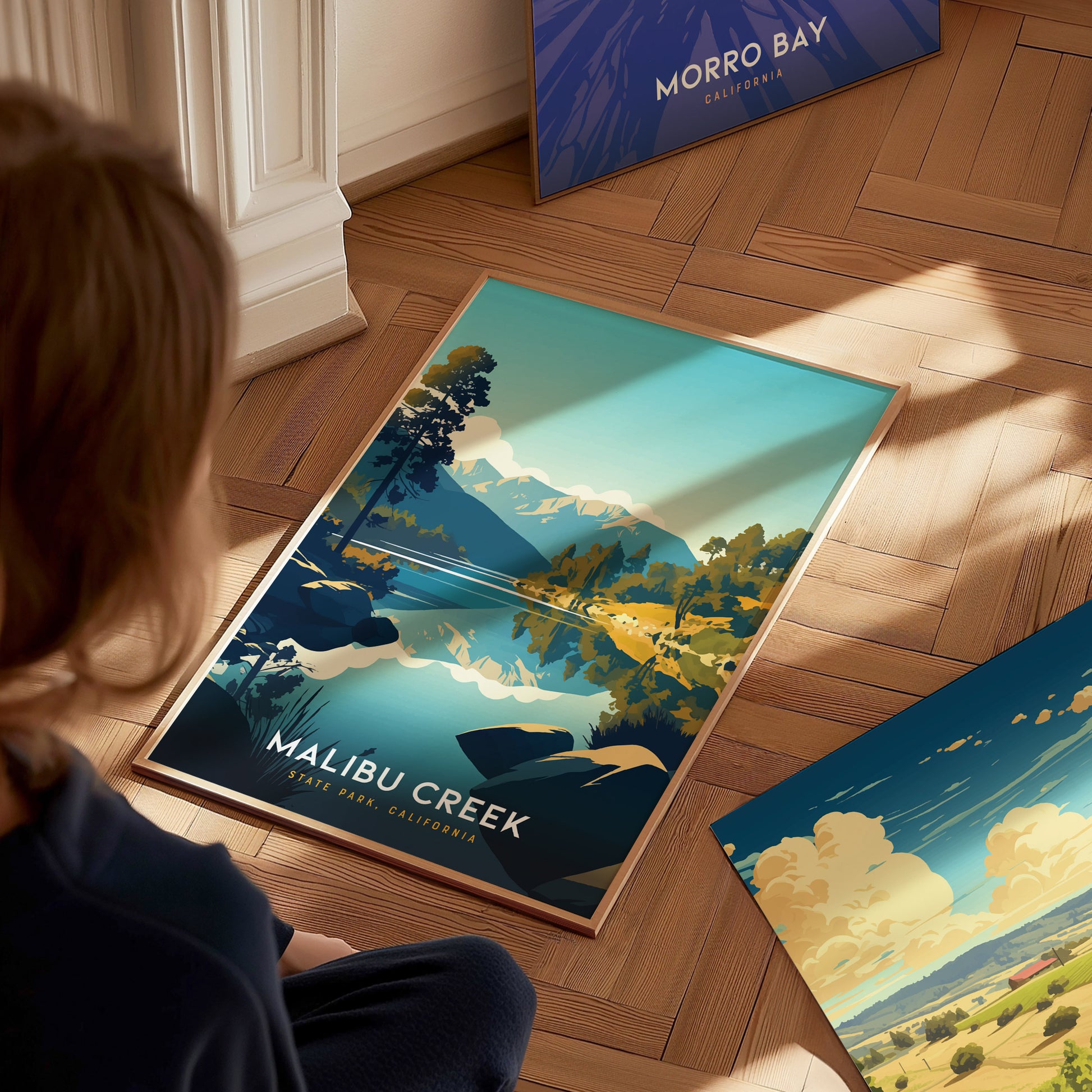 A person looks at a Malibu Creek State Park poster featuring a scenic landscape with mountains, trees, and a river. The poster rests on a wooden floor alongside another artwork.