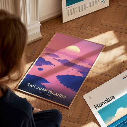 A person looks at a vibrant San Juan Islands poster on a wooden floor, featuring pink and purple tones with a setting sun. Another poster titled Honolua is partially visible.