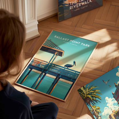 A person observes a framed poster on a wooden floor depicting Ballast Point Park with a pelican on a pier, surrounded by additional artwork.