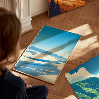 A person sits on a wooden floor, looking at a White Sands National Park poster featuring blue skies and white dunes. Other posters with landscape scenes are nearby.