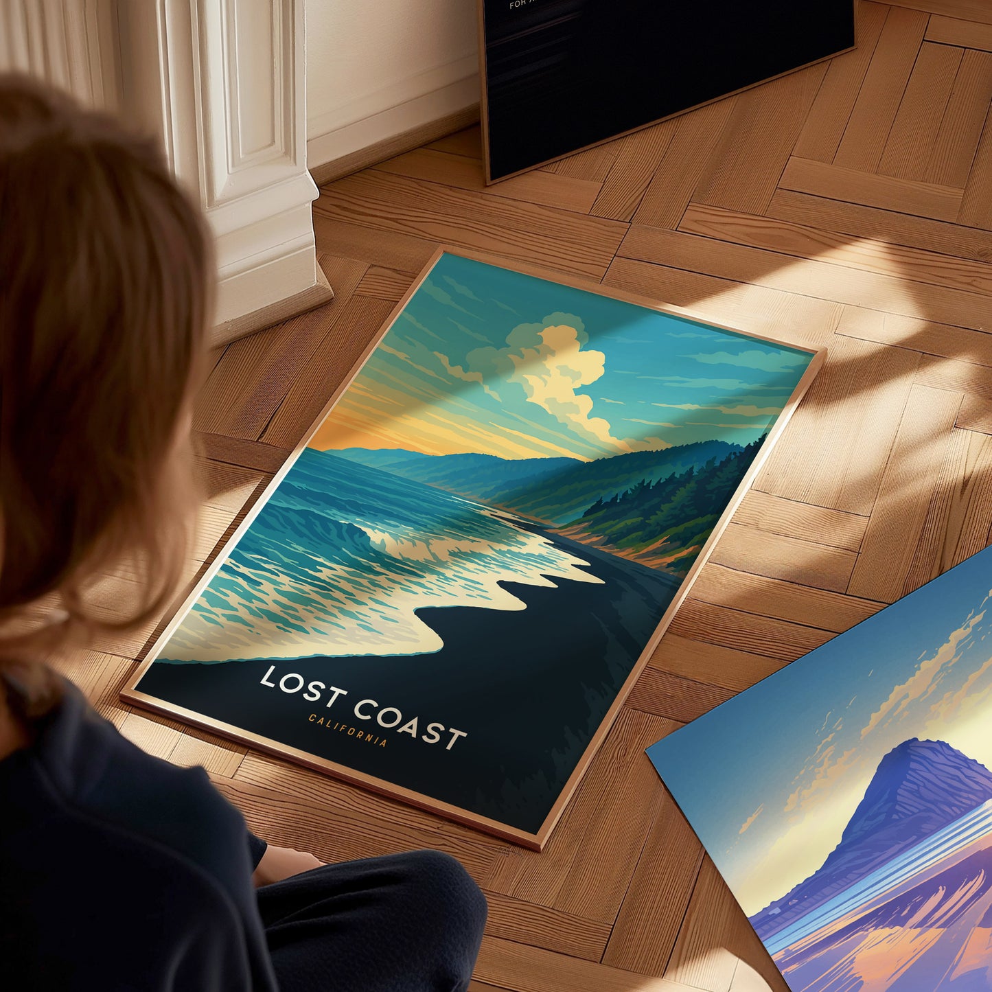 A child sits on a wooden floor, gazing at a vibrant poster titled Lost Coast California. The artwork features a serene coastal landscape with rolling waves, lush green hills, and a colorful sky at sunset, creating a peaceful ambiance.