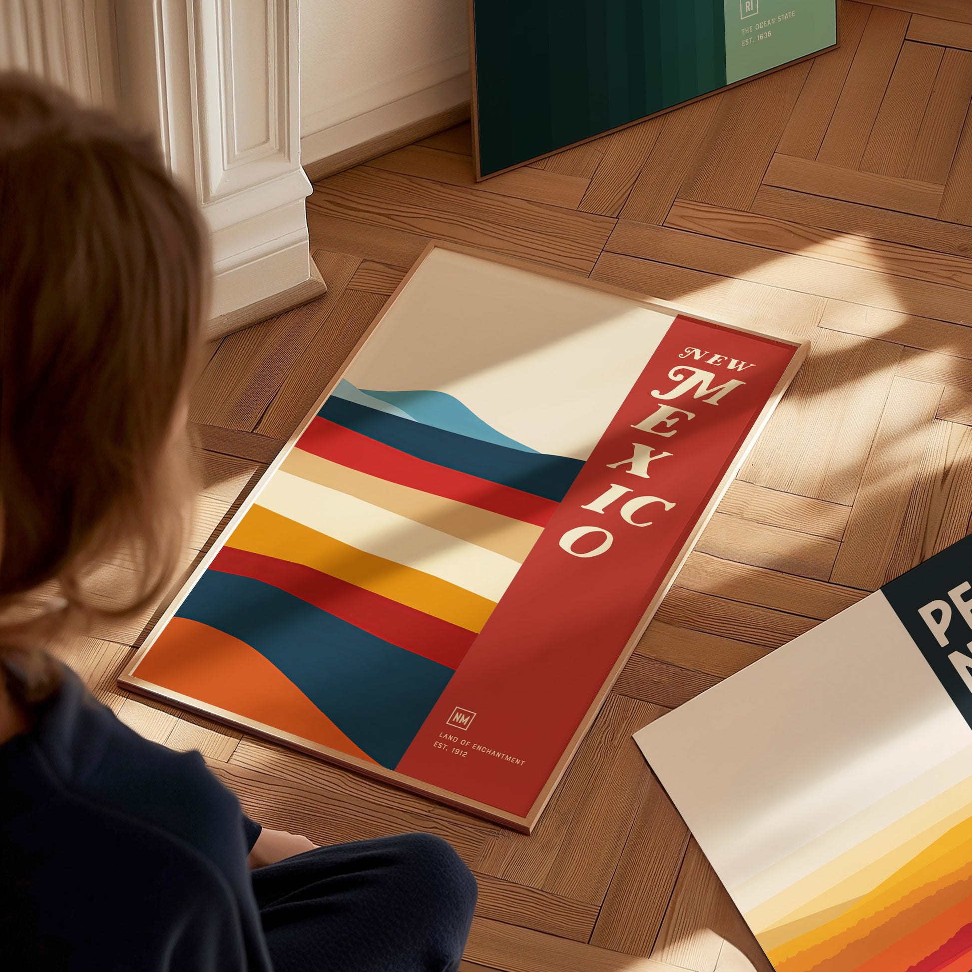 A person looks at a colorful New Mexico poster on a wooden floor, featuring abstract stripes and bold text. Sunlight casts gentle shadows across the scene.