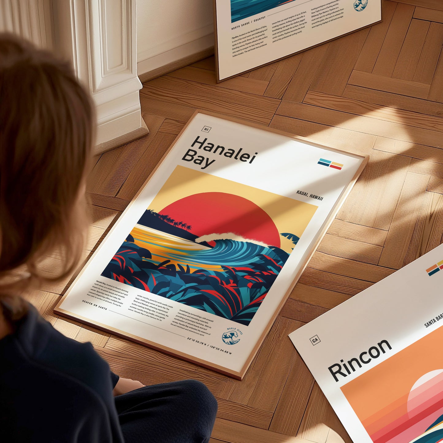 A person sits on a wooden floor, looking at a framed Hanalei Bay poster featuring a vibrant sunset, curling wave, and tropical foliage. Other posters are partially visible nearby.