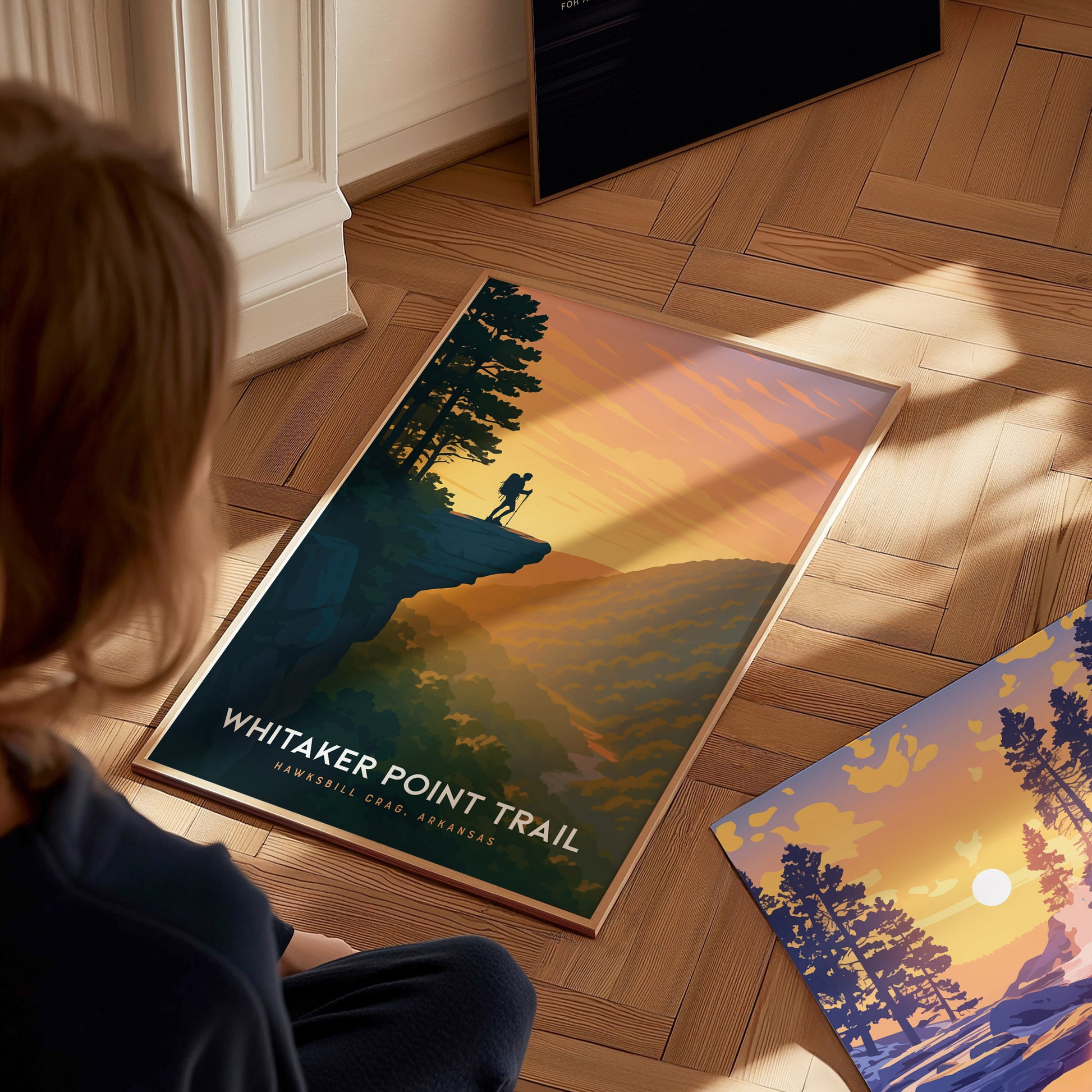 A person is sitting on a wooden floor, looking at a framed poster of Whitaker Point Trail. The poster features a scenic view of a hiker on a cliff with warm orange and yellow hues illuminating the landscape and a serene sunset in.
