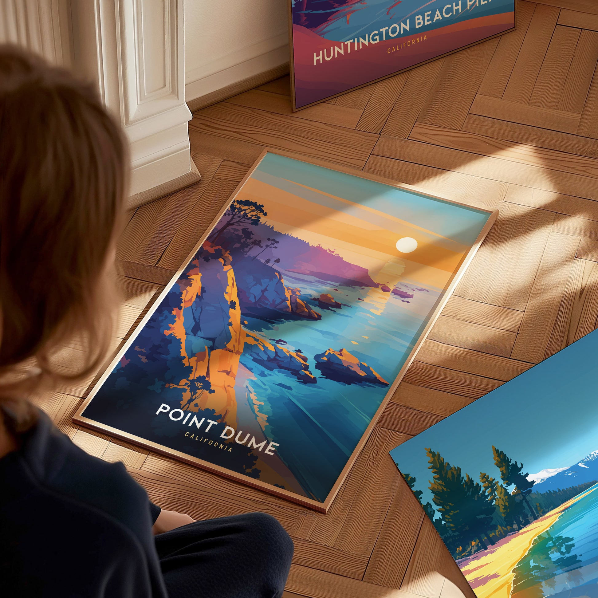 A person sits on a wooden floor, looking at a vibrant Point Dume California poster featuring cliffs, ocean, and sunset. Nearby, two other colorful posters are partially visible.