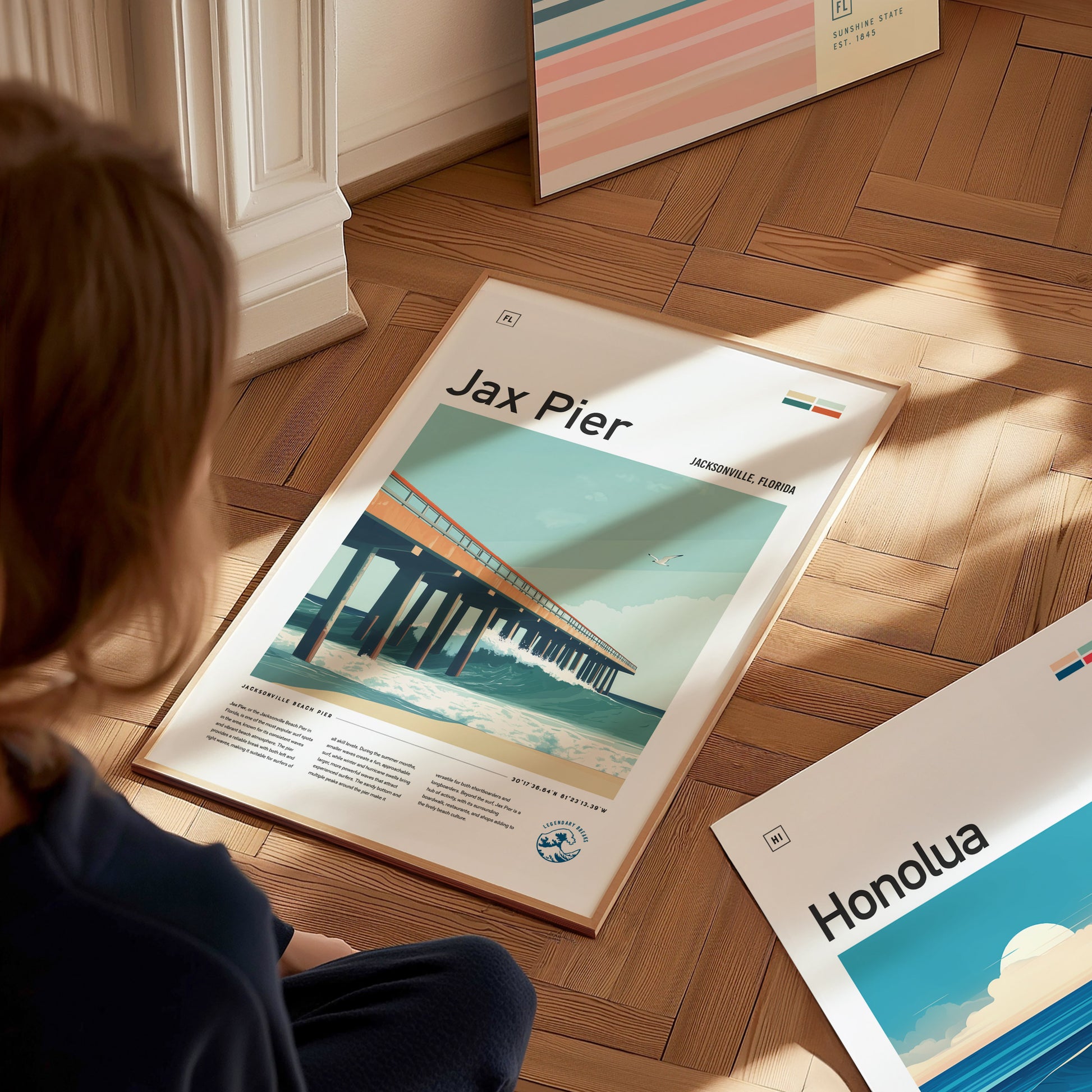 A person is seated on a wooden floor, looking at framed posters, including a Jax Pier Surf Poster featuring an ocean scene and pier. Natural light illuminates the scene.