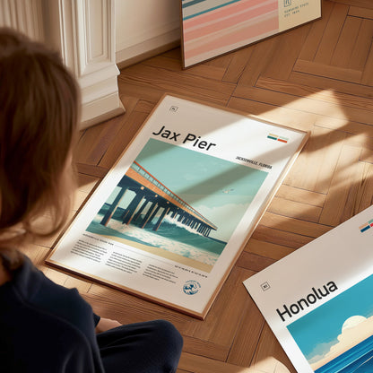 A person is seated on a wooden floor, looking at framed posters, including a Jax Pier Surf Poster featuring an ocean scene and pier. Natural light illuminates the scene.